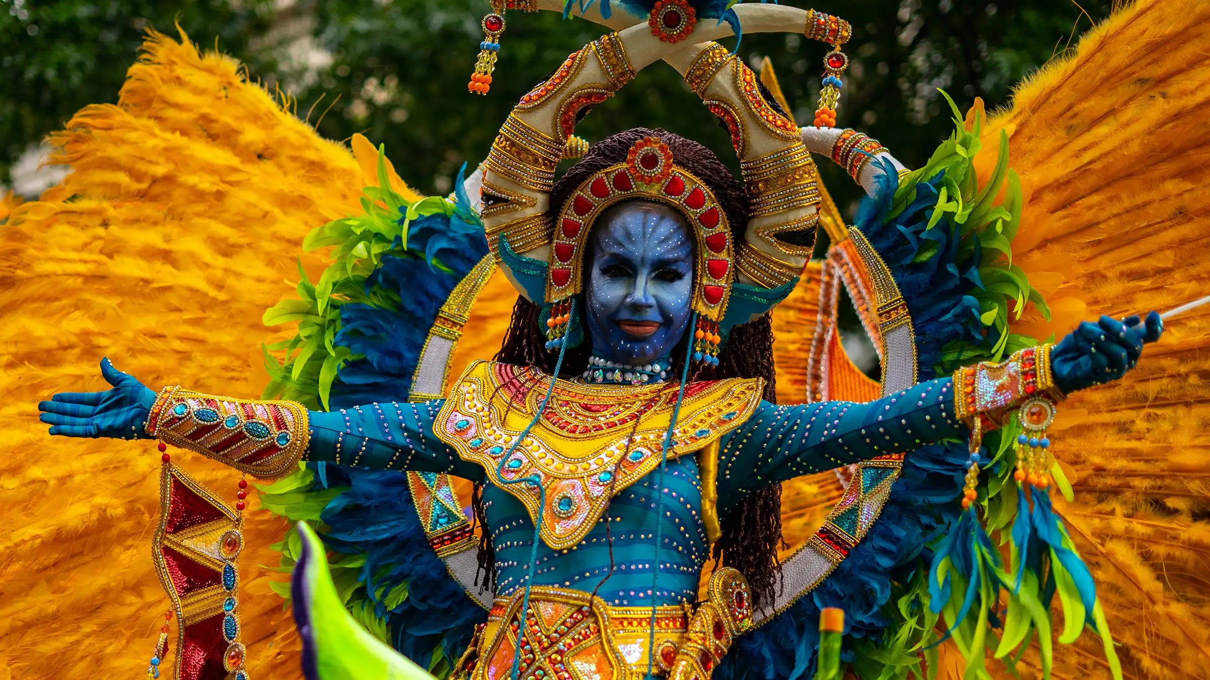 Arts-Culture-Banner-1 A vibrant performer wears an elaborate costume inspired by fantasy and cultural motifs. Their face is painted blue with intricate white markings, and they wear a beaded headdress adorned with red, gold, and turquoise details, featuring curved horn-like structures.