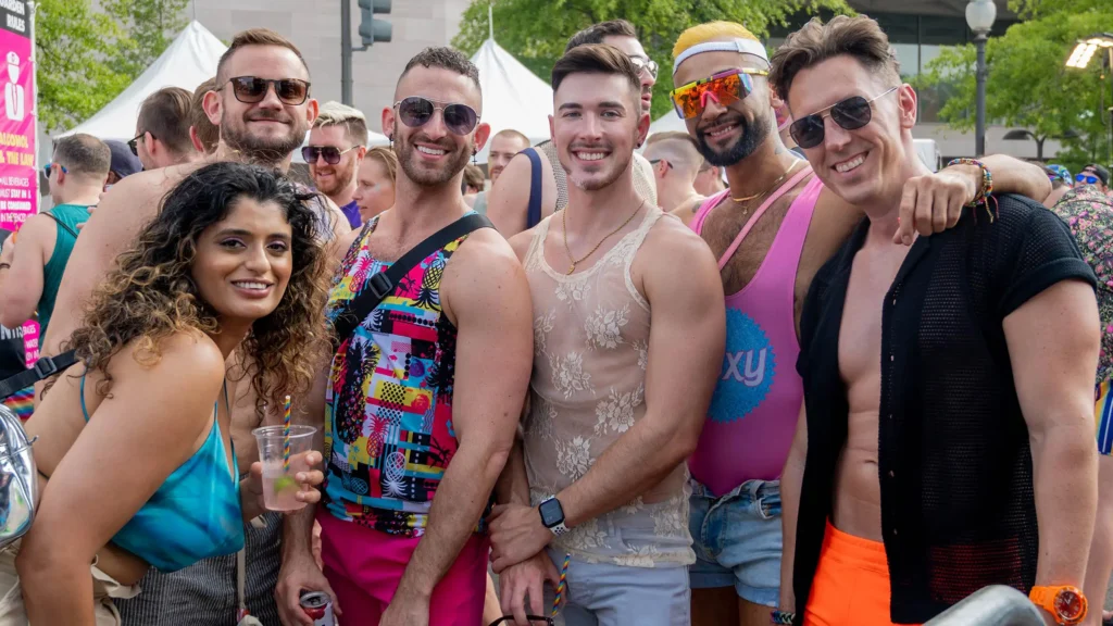 A group of people smiling at an outdoor event. They are dressed in colorful and vibrant clothing, including tank tops and sunglasses. The atmosphere appears festive, with tents and other attendees visible in the background.