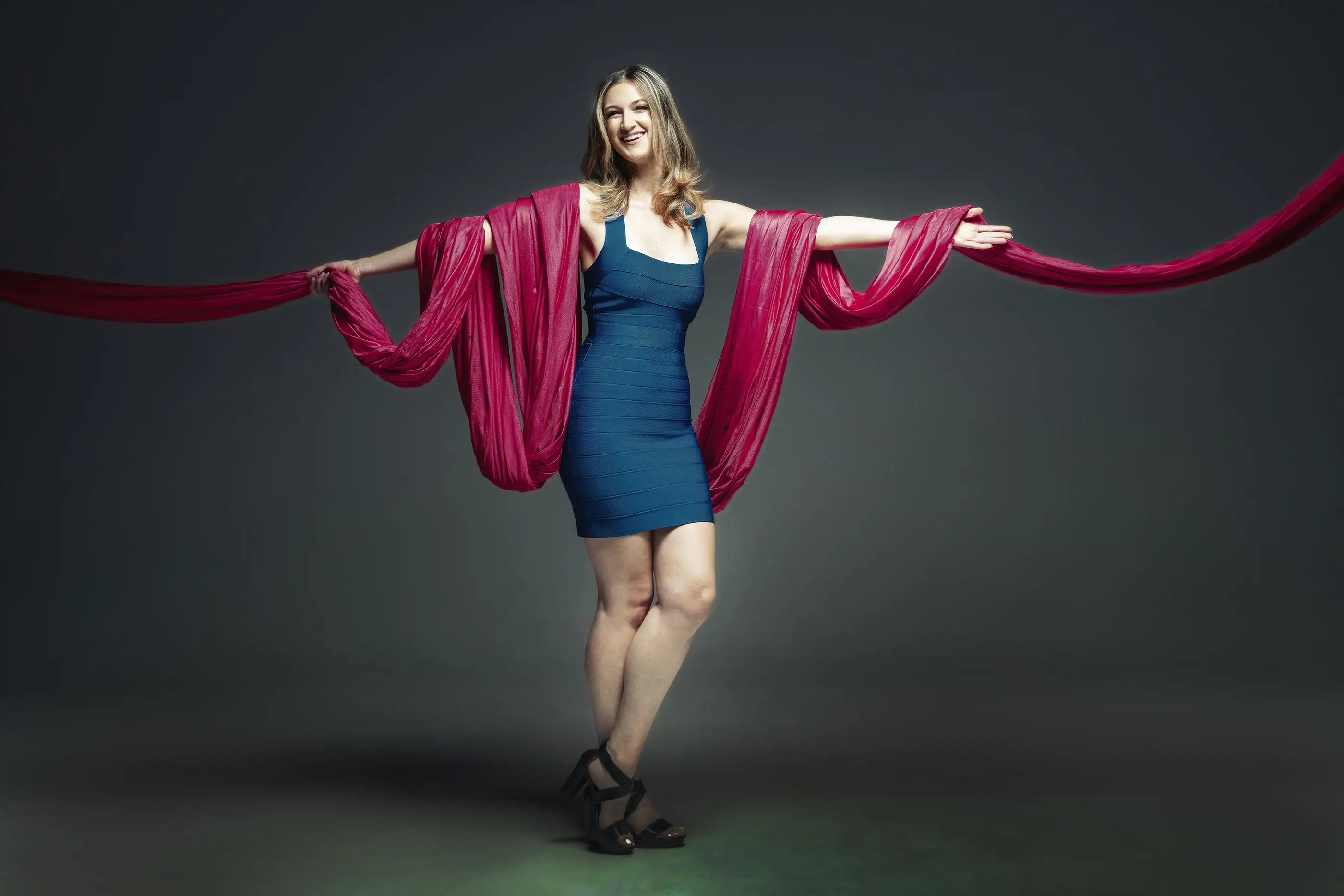 Person in a blue dress stands smiling, holding a long fuchsia fabric draped around their arms against a dark background.