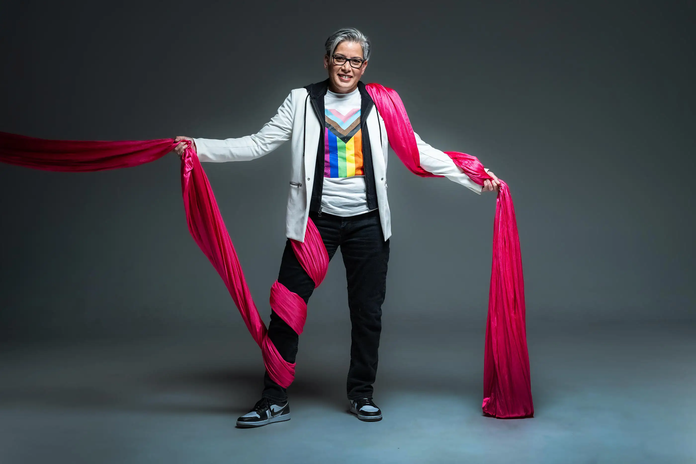 Person smiling, wrapped in flowing fuchsia fabric. They wear a white jacket, colorful rainbow shirt, black pants, and sneakers. The background is a smooth gray.