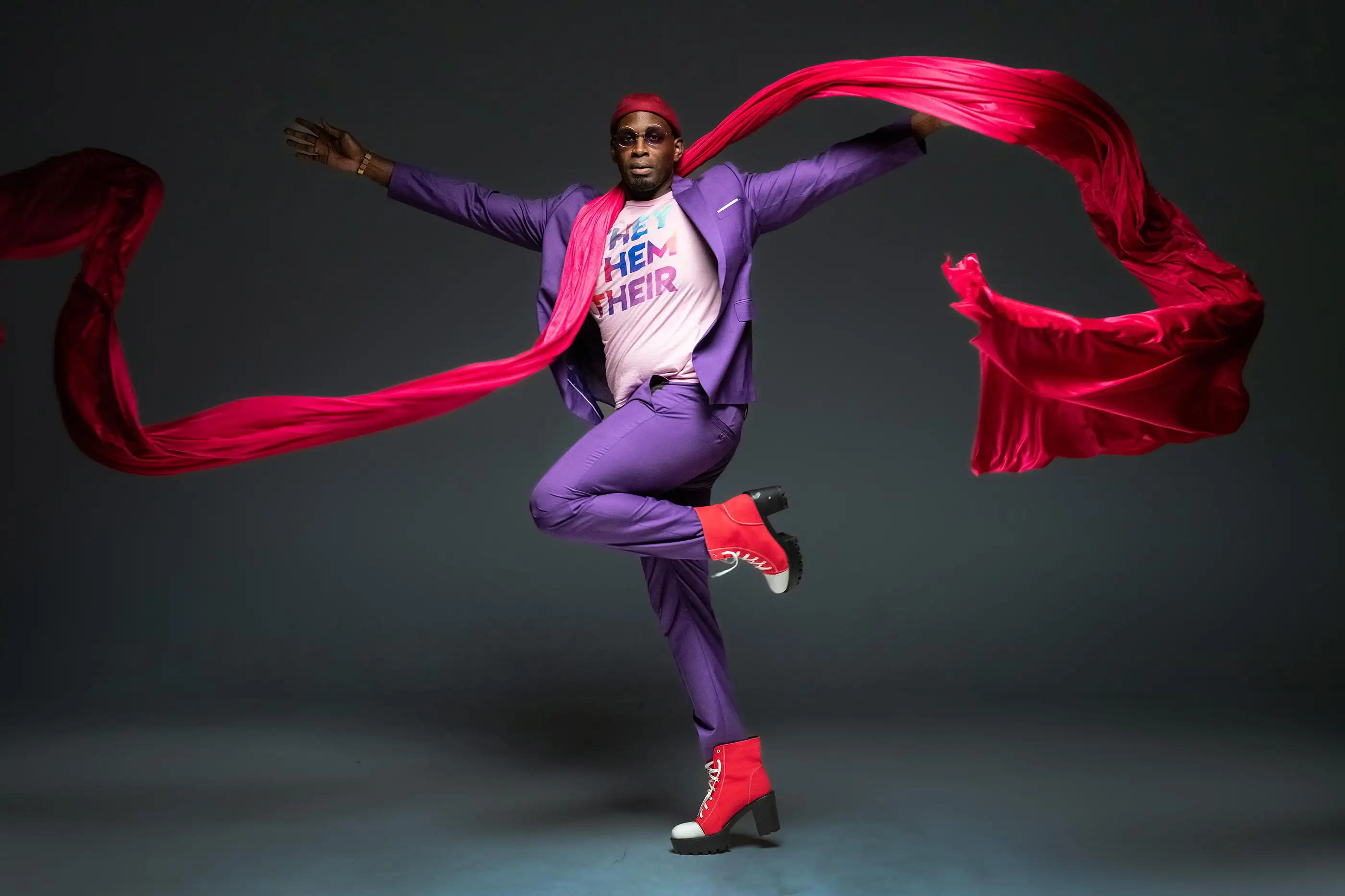A person stylishly leaps in mid-air wearing a purple suit, pink T-shirt with colorful text, pink boots. Flowing fuchsia fabric trails dramatically from their raised hands against a dark background.