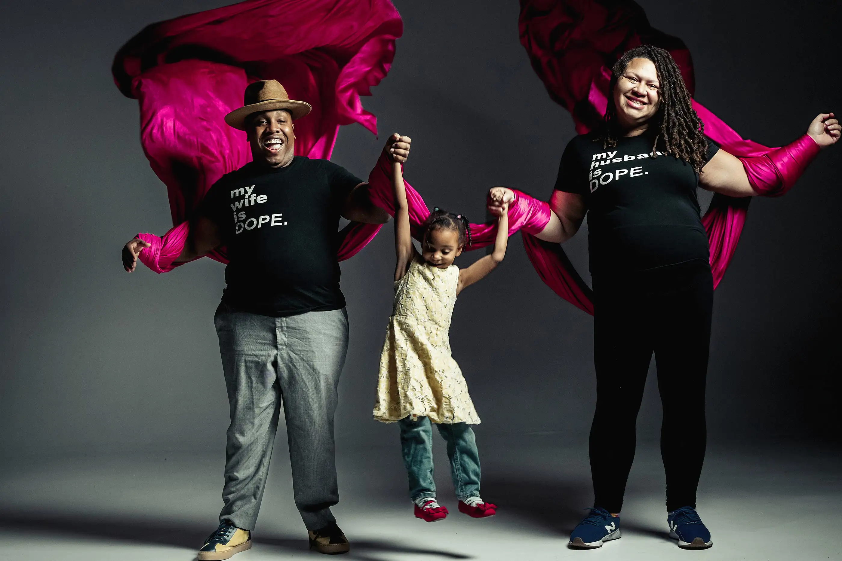 A joyful family moment: two adults hold hands with a child between them. The adults wear matching shirts with playful messages. Vibrant fuchsia fabric flows behind them. The child, smiling, is lifted slightly off the ground, wearing a yellow dress.