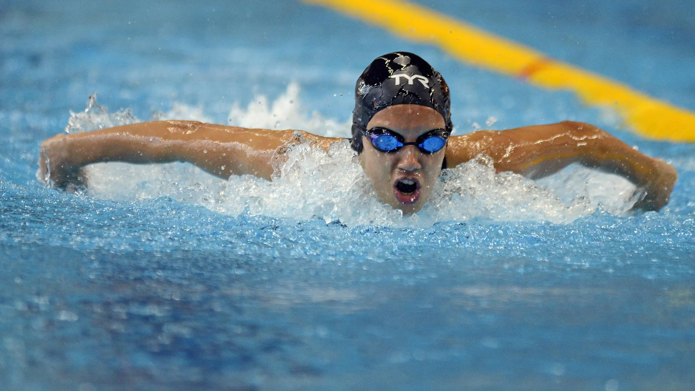Sports-Banner A swimmer, wearing a black swim cap and blue goggles, is performing the butterfly stroke in a pool.