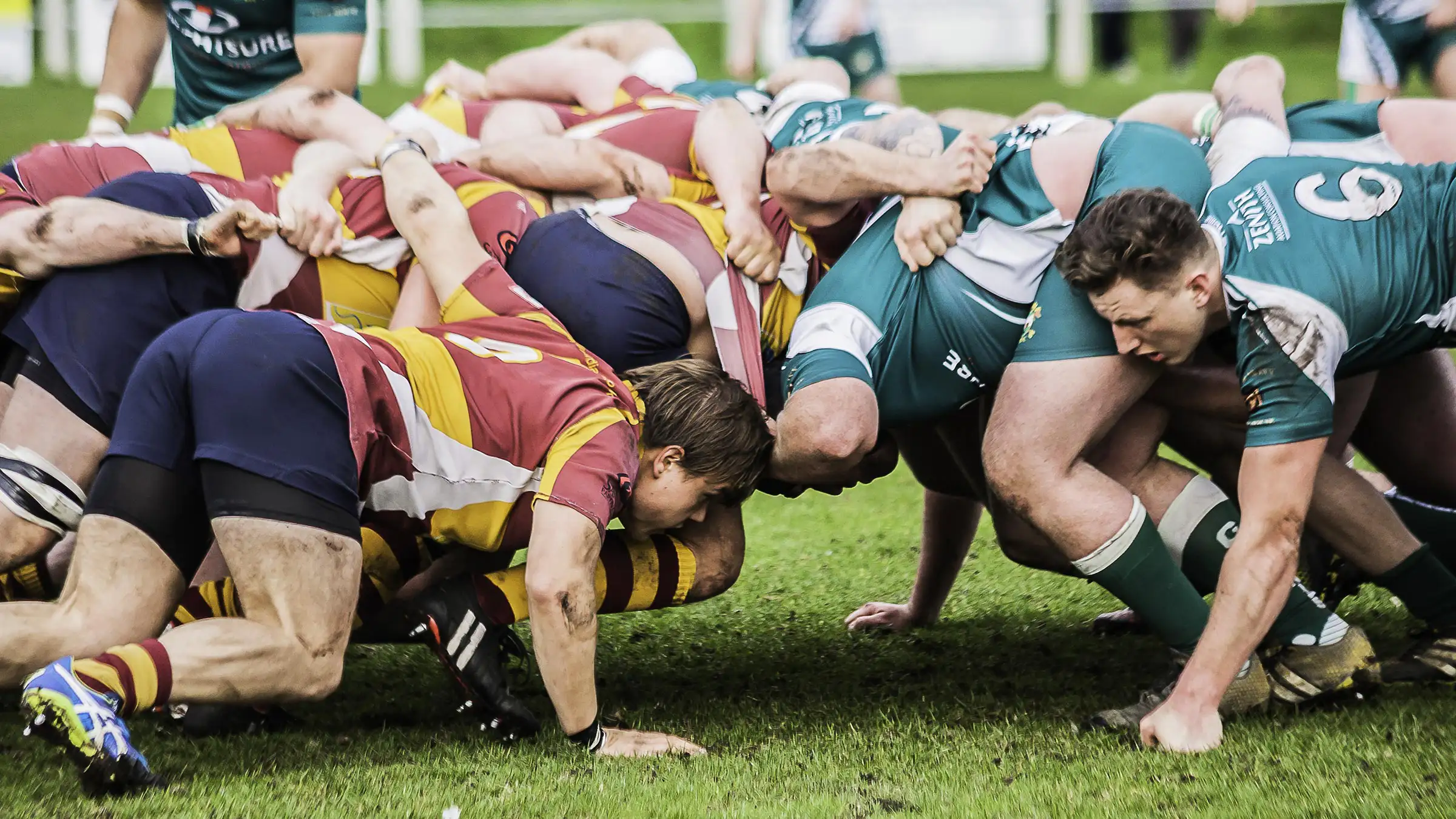 Rugby players engage in a scrum, with teams in red and yellow striped jerseys on the left and green jerseys on the right. The players are locked in formation on a grassy field during a match.