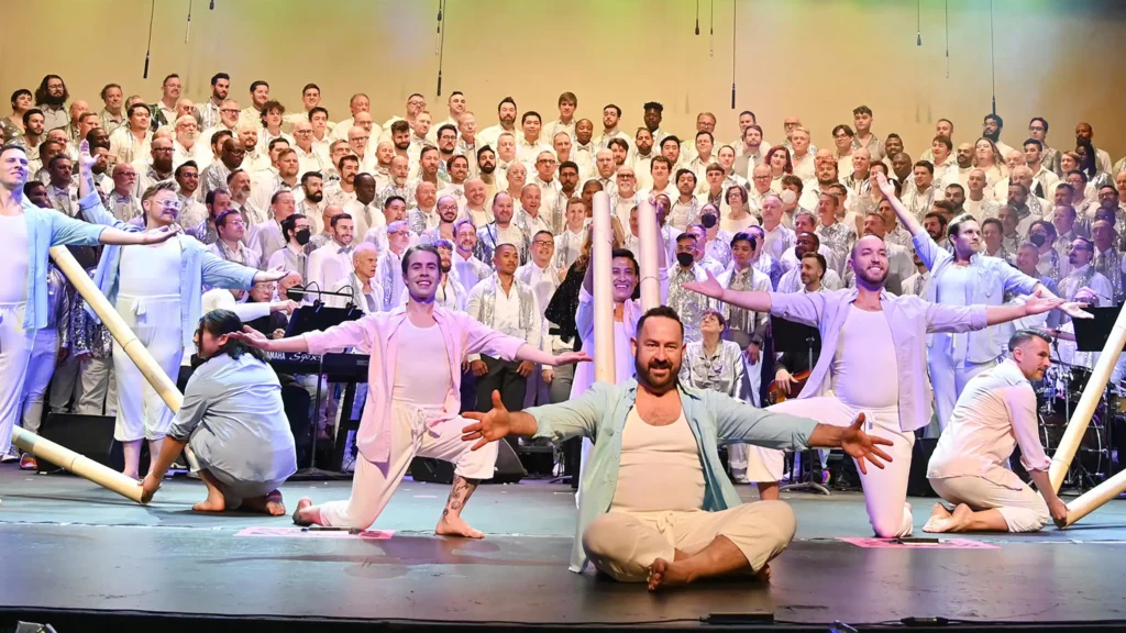 A large choir dressed in white performs on stage while a group of dancers in pastel outfits poses energetically in front. The stage is brightly lit, and musical instruments are visible in the background.