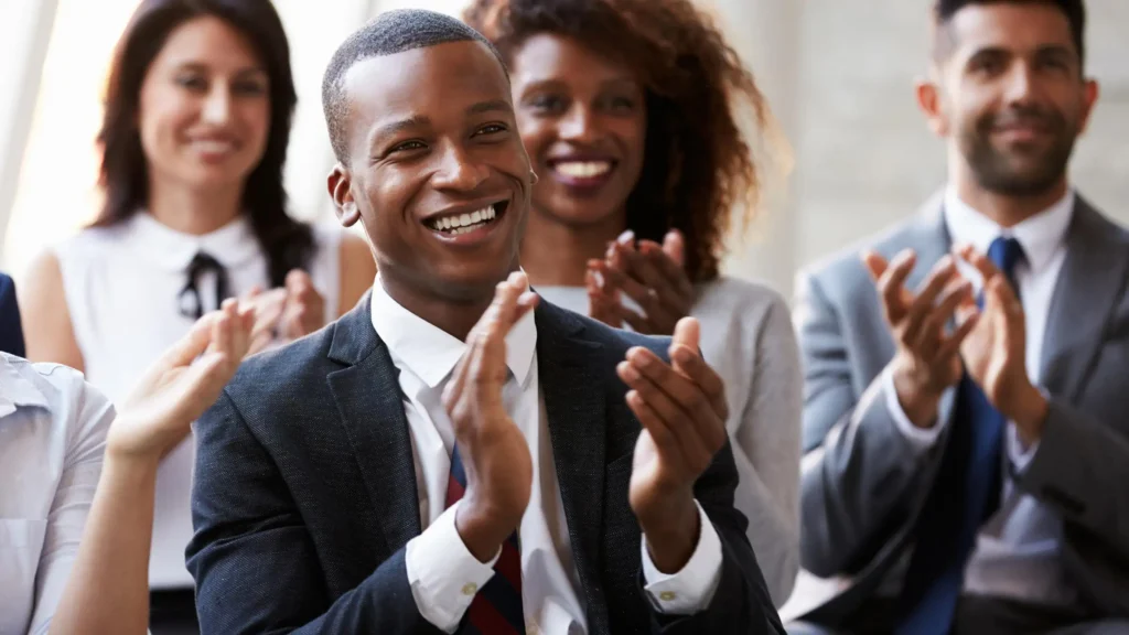 A diverse group of people in business attire sitting and clapping. The foreground shows a smiling person in a suit. Others in the background are also smiling and clapping, conveying a positive, enthusiastic atmosphere.