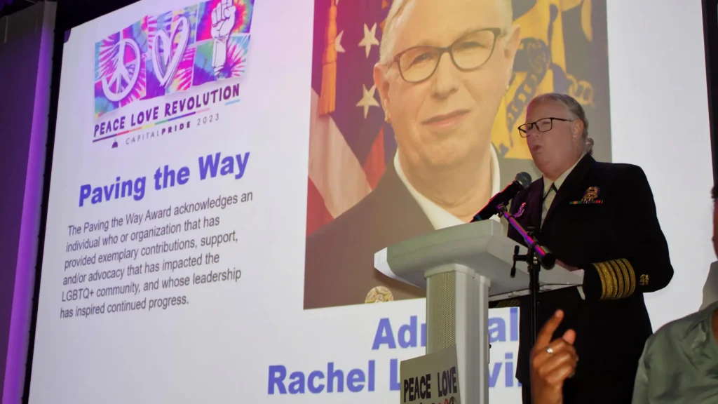 A speaker in uniform stands at a podium, addressing an audience. A large screen displays an award titled Paving the Way, featuring a photo of a person and text about the recipient. The event backdrop reads Peace Love Revolution with vibrant colors.