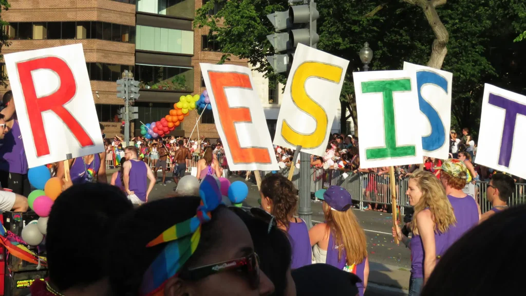 People marching holding large letters spelling RESIST in various colors. A colorful balloon arch and a crowd of spectators are visible in the background.