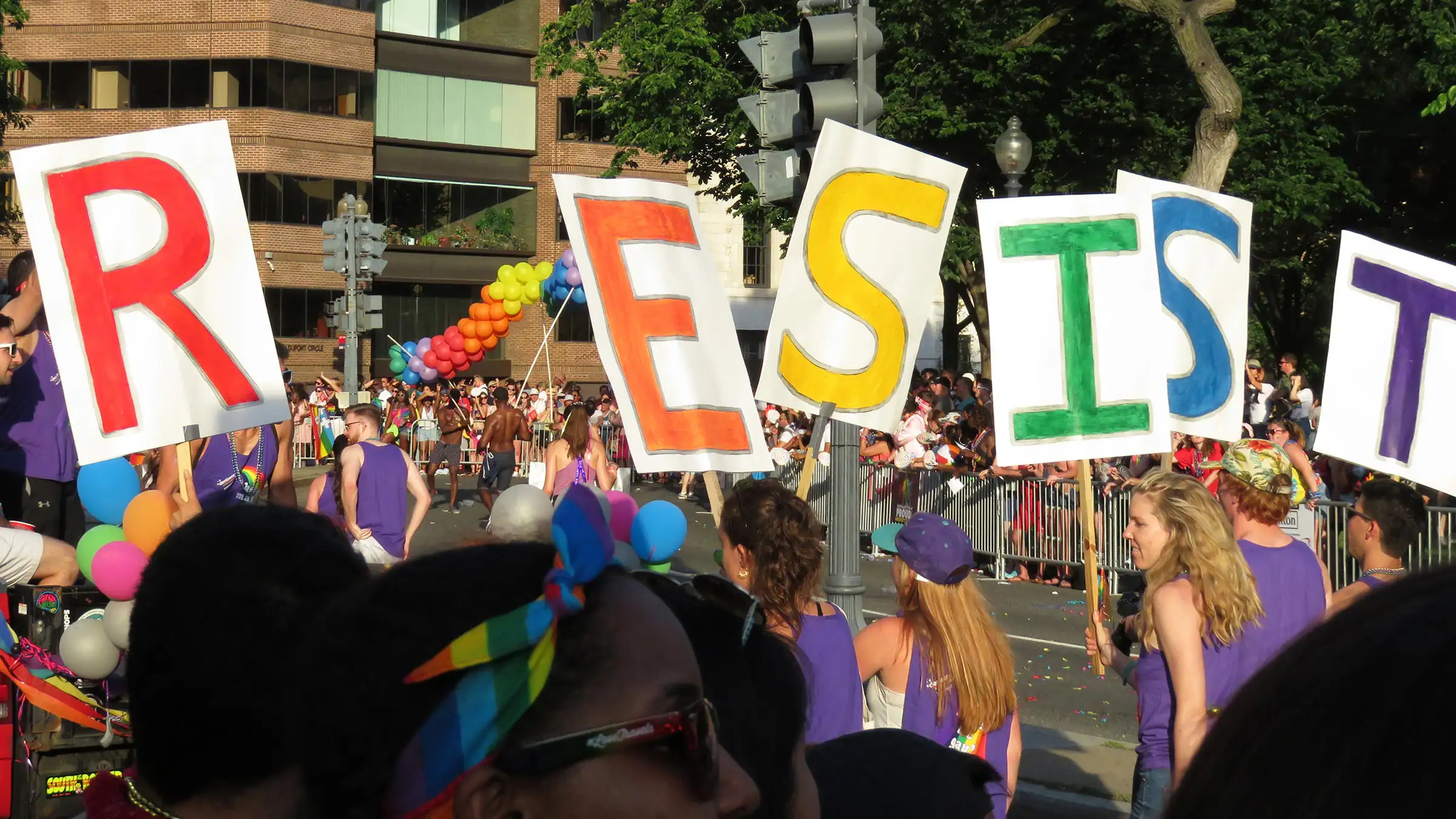 March-Banner People marching holding large letters spelling RESIST in various colors. A colorful balloon arch and a crowd of spectators are visible in the background.
