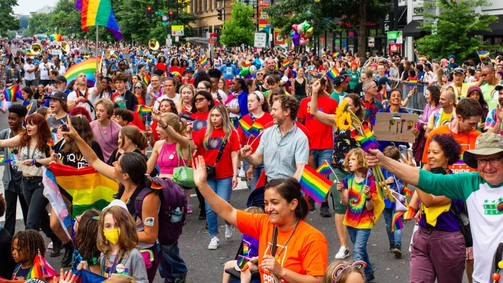 A diverse group of people joyfully participate in a street parade, waving rainbow flags. The street is filled with colorful outfits and lively expressions. Trees line the sidewalk, and buildings are visible in the background.