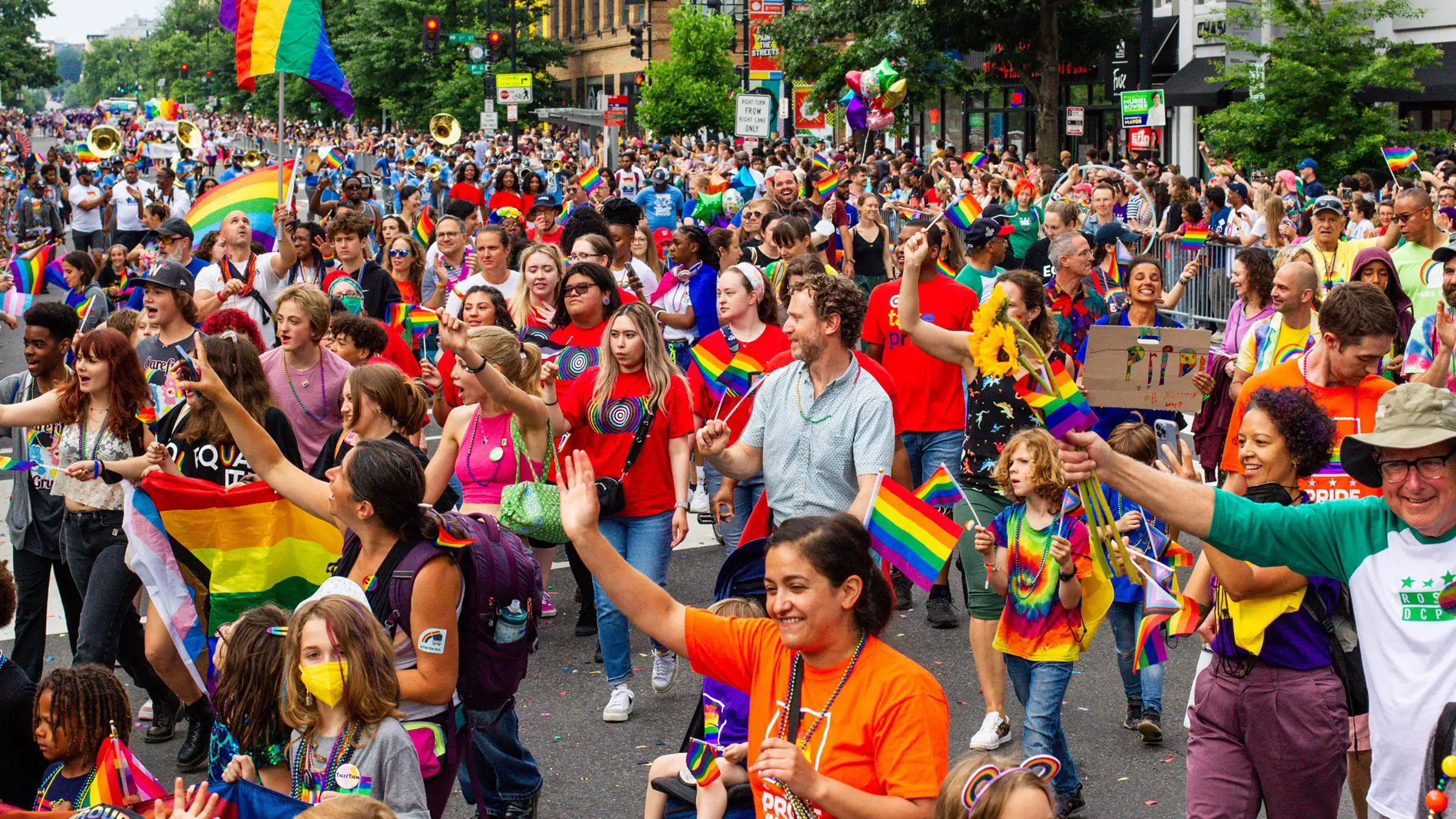 Parade-Banner A diverse group of people joyfully participate in a street parade, waving rainbow flags. The street is filled with colorful outfits and lively expressions. Trees line the sidewalk, and buildings are visible in the background.