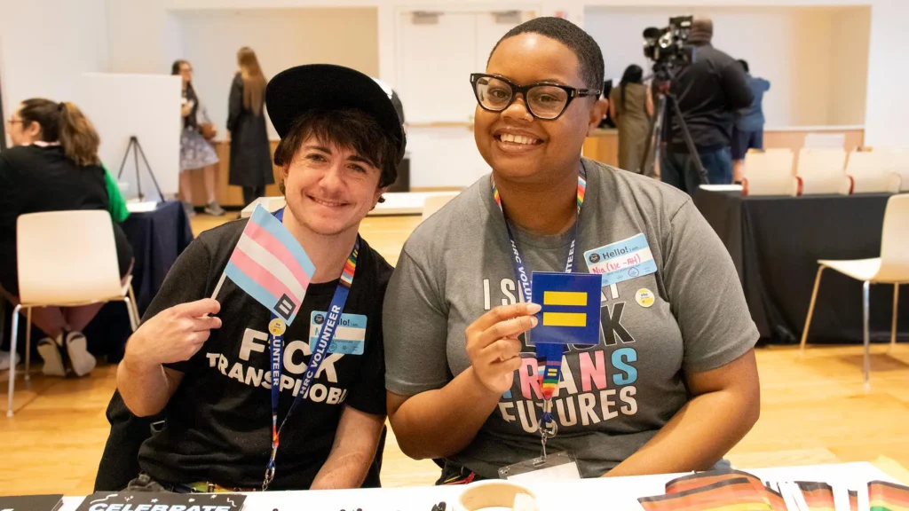 Two people smiling at a table. The person on the left holds a trans pride flag and wears a black shirt. The person on the right holds an equality symbol and wears a gray shirt.