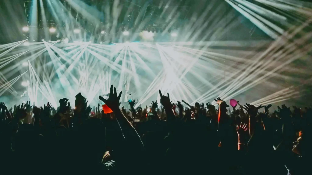Concert crowd with hands raised, illuminated by bright, colorful stage lights and beams, creating a vibrant, dynamic atmosphere.