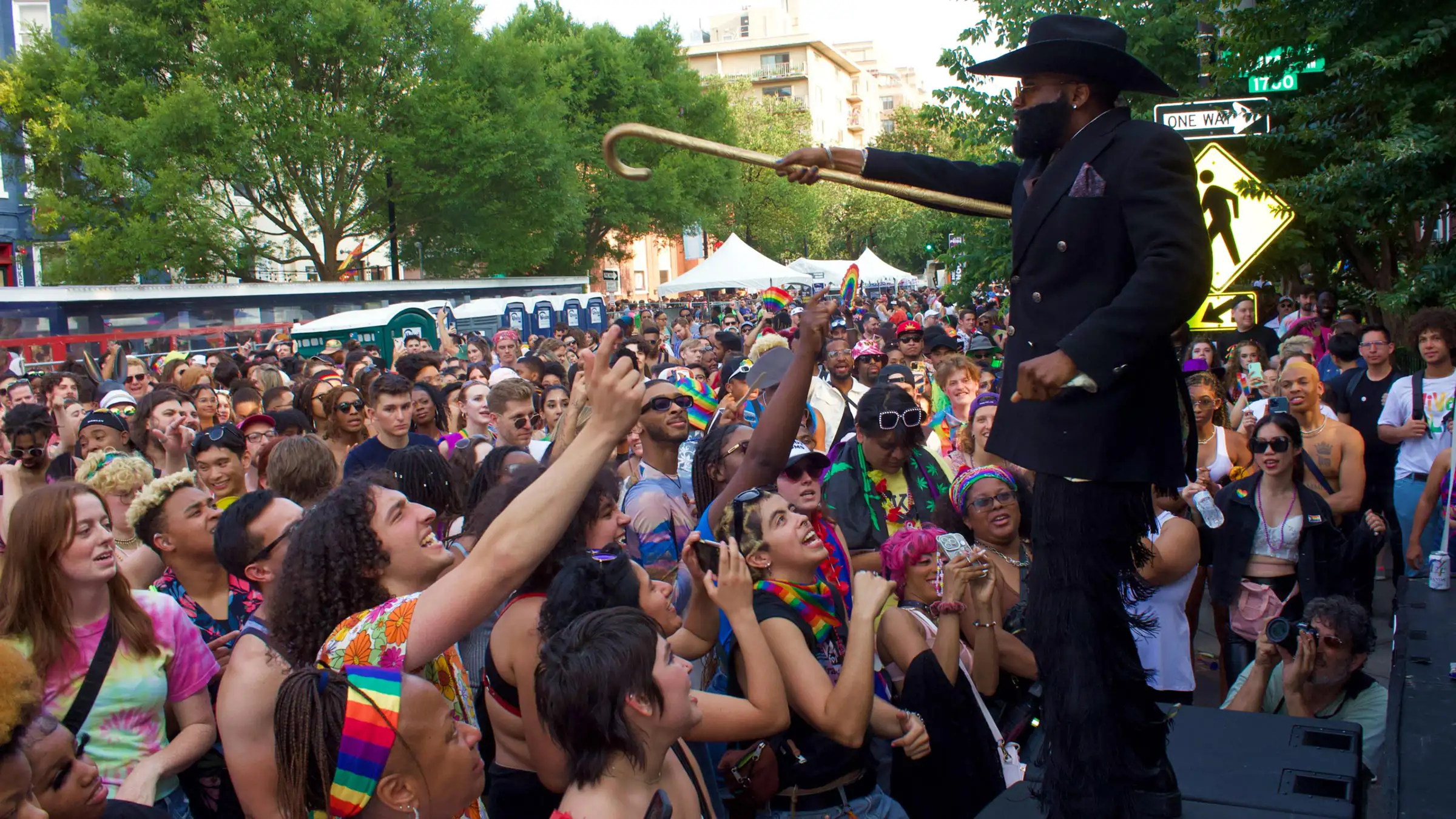 A person in a black hat and coat stands onstage holding a cane, engaging with a diverse, cheering crowd during an outdoor event. The background features trees, buildings, and tents under a clear sky.