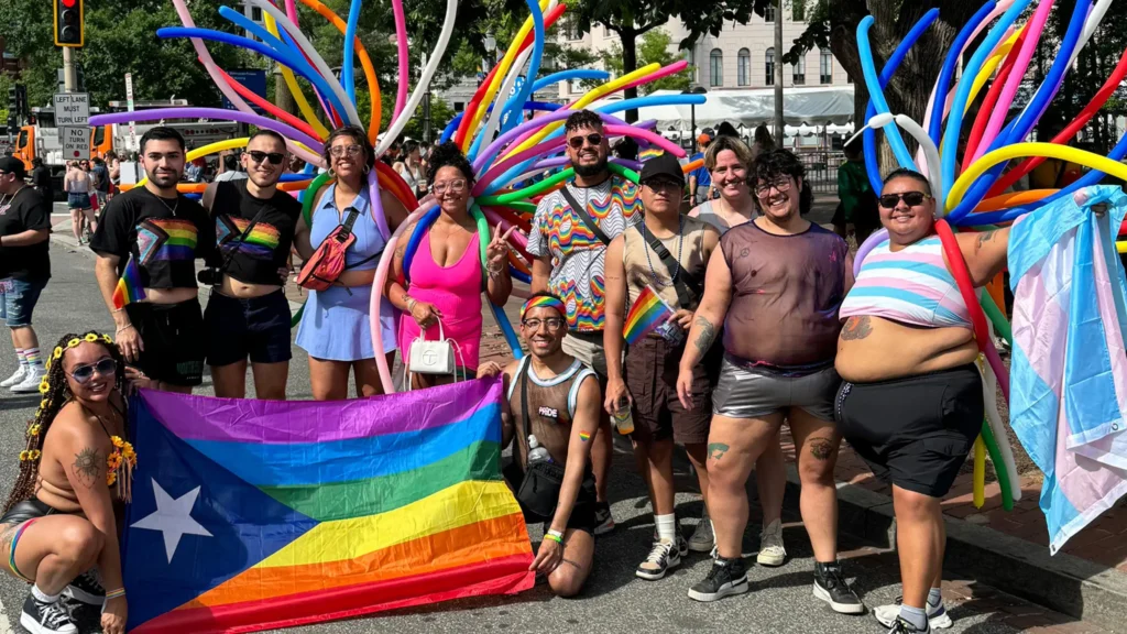 A group of diverse individuals poses together at a Pride event, dressed in colorful outfits, including rainbow-themed clothing, mesh tops, and accessories.