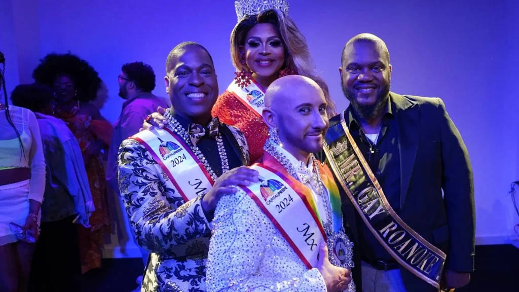Four people in a celebratory setting, with two wearing sashes. They stand together smiling, one wearing a crown.