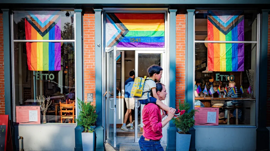 A person carries a child on their shoulders in front of a brick building with large windows displaying rainbow and Progress Pride flags. The scene is lively, with other people visible inside the establishment.